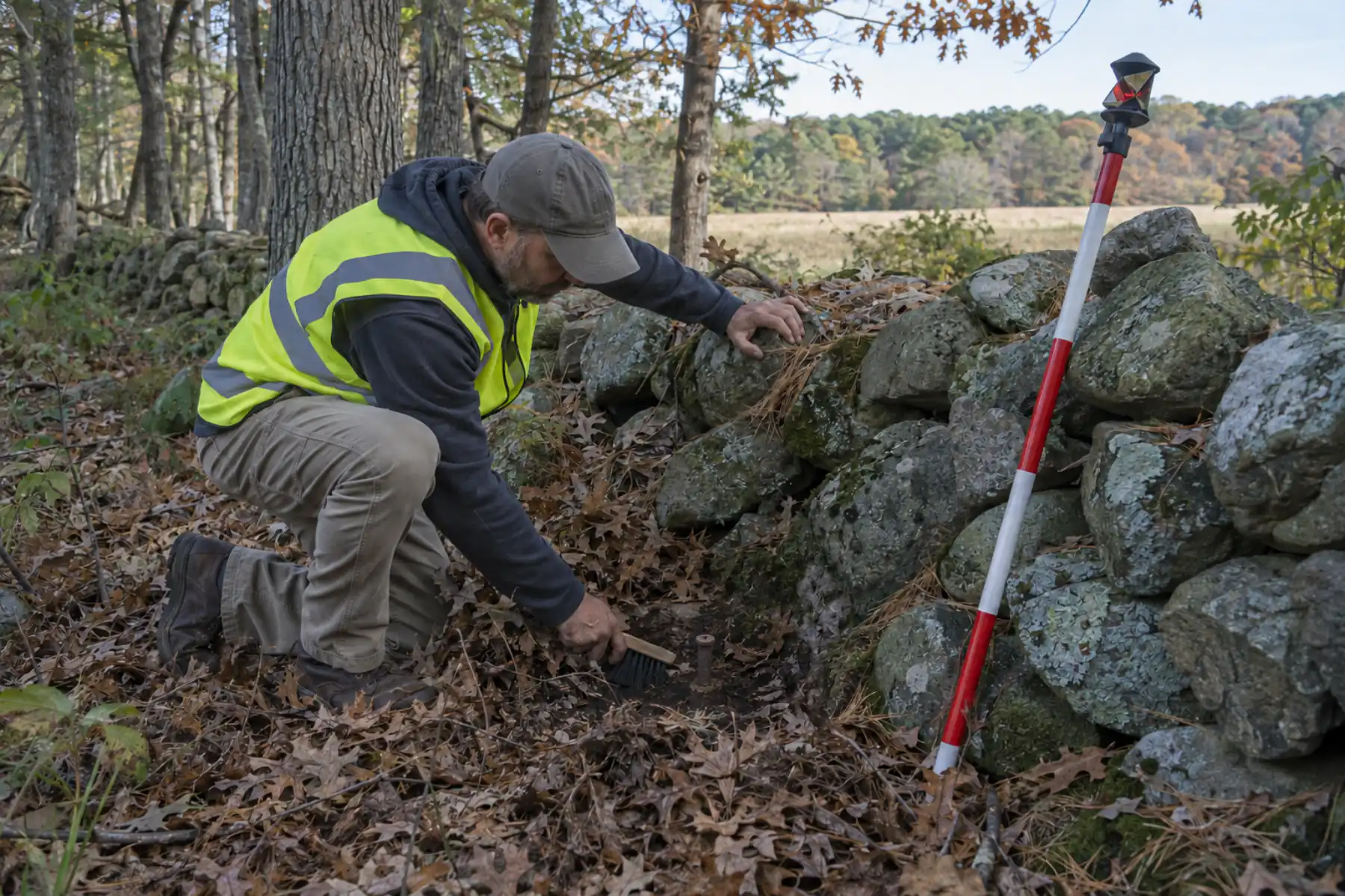 Surveyor clearing leaves from an iron pipe marker beside a stone wall on a wooded East End parcel